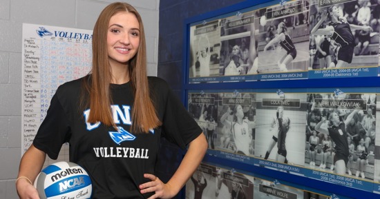 UNK sophomore Brynn Stowell poses for a photo next to the All-American display inside the volleyball locker room. Her mother, Danielle (Shum) Stowell, is the first athlete featured on the wall. (Photo by Erika Pritchard, UNK Communications)